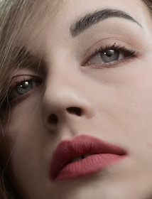 A close-up of a person's face focusing on well-defined eyebrows, expressive eyes with subtle makeup, and full lips adorned with a matte pink lipstick. The image captures fine details of the skin texture and hair strands.