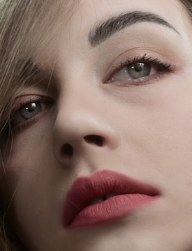 A close-up of a person's face focusing on well-defined eyebrows, expressive eyes with subtle makeup, and full lips adorned with a matte pink lipstick. The image captures fine details of the skin texture and hair strands.