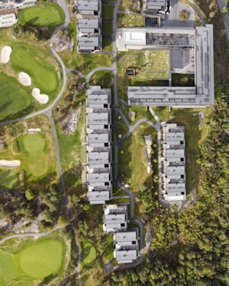 An aerial view of a modern residential complex with multiple rectangular buildings lined up parallel to each other. The buildings are surrounded by green lawns, pathways, and trees. Adjacent to the complex on the left are golf course elements, including well-maintained grass, sand bunkers, and golf holes. A pool with a clear blue color is visible on the rooftop of one of the buildings.