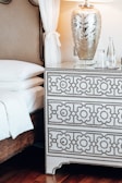 A bedroom scene featuring a bed with white linens and a decorative headboard. Next to the bed is a patterned white and black dresser with intricate designs. On top of the dresser, there's a large decorative lamp with an ornate silver base and a glass finish, creating an elegant ambiance. Beside the lamp are a few clear glass bottles and glasses. The room is softly lit, enhancing the luxurious texture of the furnishings.