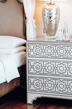 A bedroom scene featuring a bed with white linens and a decorative headboard. Next to the bed is a patterned white and black dresser with intricate designs. On top of the dresser, there's a large decorative lamp with an ornate silver base and a glass finish, creating an elegant ambiance. Beside the lamp are a few clear glass bottles and glasses. The room is softly lit, enhancing the luxurious texture of the furnishings.