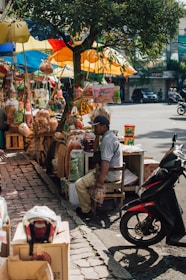 A street vendor is seated on a stool surrounded by various packaged snacks and goods hanging from a tree. Brightly colored umbrellas, predominantly yellow, blue, and red, provide shade. A motorbike is parked nearby, and there are boxes with items such as a helmet on the ground. The scene captures a casual, everyday street market atmosphere.