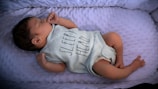 A newborn baby is lying on a soft, textured surface. The baby is wearing a light-colored onesie with the words 'Sleepy Hero' printed on it. The baby's hair is dark, and they are peacefully resting with their eyes closed.