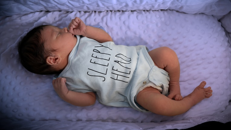 A newborn baby is lying on a soft, textured surface. The baby is wearing a light-colored onesie with the words 'Sleepy Hero' printed on it. The baby's hair is dark, and they are peacefully resting with their eyes closed.