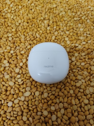 A white Realme earbud case is placed on a surface covered with numerous yellow lentils.