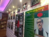 A display wall featuring photographs and framed texts. The left side has a series of portraits of various individuals, accompanied by text in a language that appears to be Bengali. On the right side, there is a large photograph of a person with glasses and additional text, alongside a flag with a red circle on a green background. The setting is well-lit, with spotlights and a purple illuminated ceiling, and features wooden shelving with books.