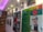 A display wall featuring photographs and framed texts. The left side has a series of portraits of various individuals, accompanied by text in a language that appears to be Bengali. On the right side, there is a large photograph of a person with glasses and additional text, alongside a flag with a red circle on a green background. The setting is well-lit, with spotlights and a purple illuminated ceiling, and features wooden shelving with books.