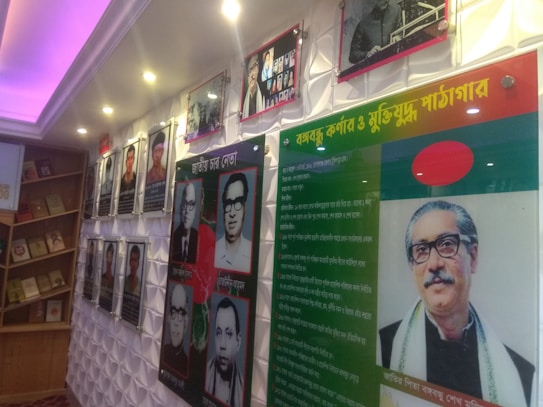A display wall featuring photographs and framed texts. The left side has a series of portraits of various individuals, accompanied by text in a language that appears to be Bengali. On the right side, there is a large photograph of a person with glasses and additional text, alongside a flag with a red circle on a green background. The setting is well-lit, with spotlights and a purple illuminated ceiling, and features wooden shelving with books.