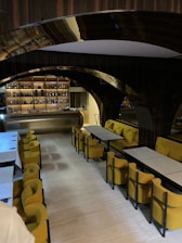 A modern, stylish interior with a bar in the background, featuring shelves stocked with various bottles. In the foreground, there are tables with yellow upholstered chairs, arranged neatly in a well-lit space. Arched design elements and a warm, inviting atmosphere are emphasized by the lighting.