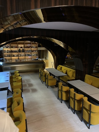 A modern, stylish interior with a bar in the background, featuring shelves stocked with various bottles. In the foreground, there are tables with yellow upholstered chairs, arranged neatly in a well-lit space. Arched design elements and a warm, inviting atmosphere are emphasized by the lighting.