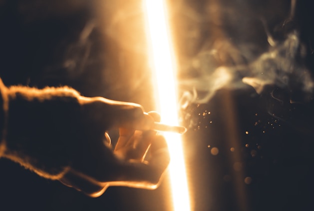 A hand is extended holding a cigarette against a bright, glowing light. Smoke swirls around, creating a hazy atmosphere. Particles appear to be floating in the air, adding to the moodiness of the scene.
