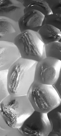 A close-up view of a textured surface with a pattern resembling hexagonal cells. The surface appears metallic or glass-like, with each cell having a smooth, reflective surface that catches light, creating a range of highlights and shadows.