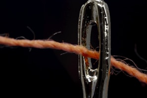 A close-up view of a sewing needle with a thread passing through its eye. The needle is metallic and shiny, and the thread is orange, showing fine details of the fibers. The background is dark, creating a strong contrast with the metallic sheen of the needle.