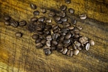 A pile of roasted coffee beans scattered on a textured wooden surface. The rich, brown beans have visible seams and a shiny appearance, indicating freshness.