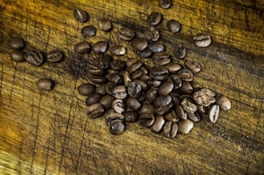 A pile of roasted coffee beans scattered on a textured wooden surface. The rich, brown beans have visible seams and a shiny appearance, indicating freshness.