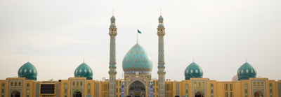 A large mosque with a central blue dome and multiple smaller domes on each side. Two tall minarets flank the central structure, adorned with intricate patterns. The architecture features a combination of yellow and turquoise tiles, showcasing detailed craftsmanship.