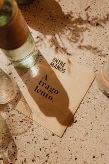 A tabletop scene with a beige paper card displaying text 'A trago lento.' and 'ENTRE MANOS.' The paper rests on a speckled surface with scattered shadows and a few partially visible bottles, suggesting a relaxed and casual setting.