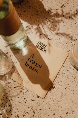 A tabletop scene with a beige paper card displaying text 'A trago lento.' and 'ENTRE MANOS.' The paper rests on a speckled surface with scattered shadows and a few partially visible bottles, suggesting a relaxed and casual setting.