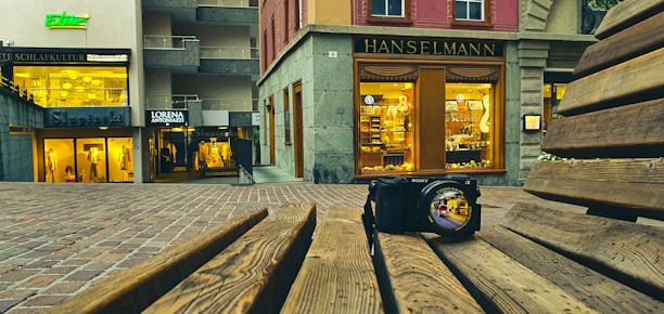A digital camera sits on a wooden bench with the camera lens reflecting a vibrant urban scene. In the background, there are various storefronts with illuminated signs and displays, including 'Hanselmann' and 'Lorena Antoniazzi'. The ground is paved with bricks, and the setting appears to be in a shopping district.