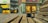 A digital camera sits on a wooden bench with the camera lens reflecting a vibrant urban scene. In the background, there are various storefronts with illuminated signs and displays, including 'Hanselmann' and 'Lorena Antoniazzi'. The ground is paved with bricks, and the setting appears to be in a shopping district.