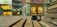 A digital camera sits on a wooden bench with the camera lens reflecting a vibrant urban scene. In the background, there are various storefronts with illuminated signs and displays, including 'Hanselmann' and 'Lorena Antoniazzi'. The ground is paved with bricks, and the setting appears to be in a shopping district.