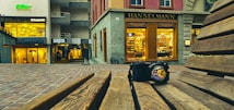 A digital camera sits on a wooden bench with the camera lens reflecting a vibrant urban scene. In the background, there are various storefronts with illuminated signs and displays, including 'Hanselmann' and 'Lorena Antoniazzi'. The ground is paved with bricks, and the setting appears to be in a shopping district.