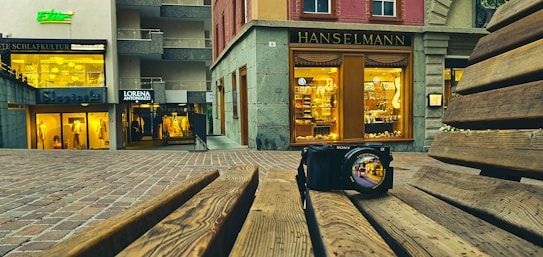 A digital camera sits on a wooden bench with the camera lens reflecting a vibrant urban scene. In the background, there are various storefronts with illuminated signs and displays, including 