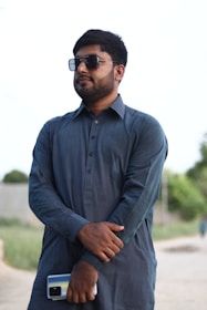 A man stands outdoors wearing sunglasses and a traditional blue kurta. He holds a smartphone in his right hand. The background features a blurry outdoor environment with greenery and a pathway.