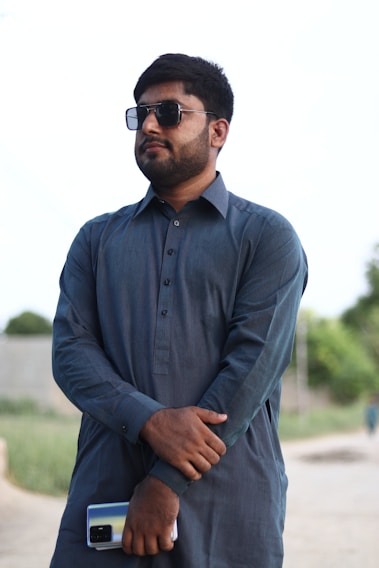 A smiling man wearing a stylish brown kurta standing in a sunlit outdoor market.