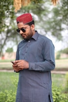 A man standing outdoors, wearing a traditional blue kurta and a red cap with sunglasses, looking at his phone in a serene, green park environment.