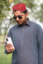 A man wearing a traditional red cap and sunglasses is looking at a smartphone. He is dressed in a dark blue kurta, standing outdoors with blurred greenery in the background.