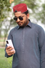 A man wearing a traditional red cap and sunglasses is looking at a smartphone. He is dressed in a dark blue kurta, standing outdoors with blurred greenery in the background.