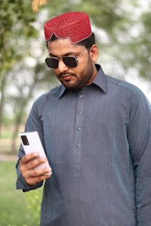 A man wearing a traditional red cap and sunglasses is looking at a smartphone. He is dressed in a dark blue kurta, standing outdoors with blurred greenery in the background.