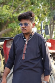 A man wearing sunglasses and a dark traditional kurta stands in front of a tractor, with green foliage visible in the background.