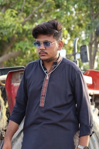 A man wearing sunglasses and a dark traditional kurta stands in front of a tractor, with green foliage visible in the background.