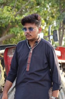 A man wearing sunglasses and a dark traditional kurta stands in front of a tractor, with green foliage visible in the background.