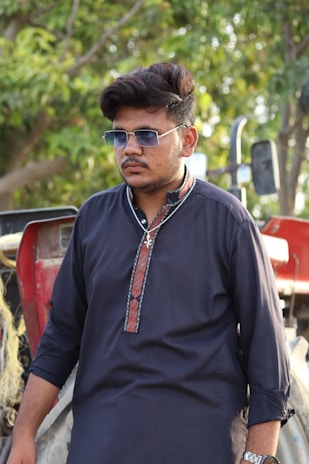 A man wearing sunglasses and a dark traditional kurta stands in front of a tractor, with green foliage visible in the background.