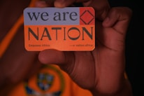 A hand is holding a card with text that reads 'we are NATION'. The card includes a red square symbol and additional text 'Empower Africa.' and a URL 'nation.africa'. The background features an orange and dark pattern.