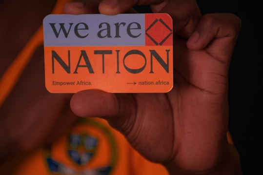 A hand is holding a card with text that reads 'we are NATION'. The card includes a red square symbol and additional text 'Empower Africa.' and a URL 'nation.africa'. The background features an orange and dark pattern.