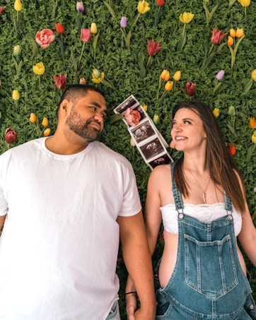 A couple is lying on a green leafy background with colorful tulips scattered around. The man, wearing a white T-shirt, and the woman in a white top and denim overalls are holding hands. Several ultrasound photos are placed between them, suggesting a pregnancy announcement.