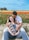 A couple sits in a field of vibrant tulips, with the woman smiling while holding ultrasound images and the man lovingly embracing her from behind. The flowers create a colorful backdrop with shades of red, orange, and yellow, and a clear blue sky stretches above.