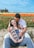 A couple sits in a field of vibrant tulips, with the woman smiling while holding ultrasound images and the man lovingly embracing her from behind. The flowers create a colorful backdrop with shades of red, orange, and yellow, and a clear blue sky stretches above.