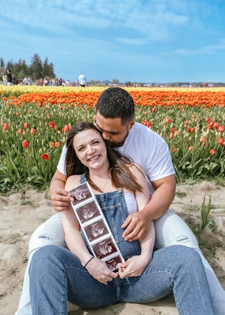A couple sits in a field of vibrant tulips, with the woman smiling while holding ultrasound images and the man lovingly embracing her from behind. The flowers create a colorful backdrop with shades of red, orange, and yellow, and a clear blue sky stretches above.