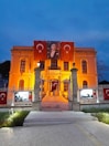 An illuminated building with an orange-yellow glow, adorned with Turkish flags. A large portrait is hung in the center, flanked by two flags featuring the crescent and star symbol. The entrance is framed by lit lamp posts and a decorative fence. The sky is a deep, twilight blue.