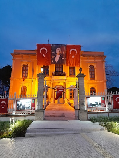 An illuminated building with an orange-yellow glow, adorned with Turkish flags. A large portrait is hung in the center, flanked by two flags featuring the crescent and star symbol. The entrance is framed by lit lamp posts and a decorative fence. The sky is a deep, twilight blue.
