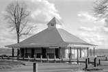 A pavilion-style structure with a pointed roof stands near a body of water. The sign reads 'Beach House,' indicating its function. The scene includes a barren tree to the left and some fluffy clouds in the sky, suggesting a slightly windy or cool day. The building is surrounded by a paved area with wooden posts and a grassy area.