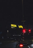 A night scene featuring an illuminated IKEA store sign in yellow with Arabic script next to it. The foreground features the rear of a vehicle with its brake lights glowing red, adding contrast against the dark surroundings.