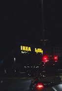 A night scene featuring an illuminated IKEA store sign in yellow with Arabic script next to it. The foreground features the rear of a vehicle with its brake lights glowing red, adding contrast against the dark surroundings.