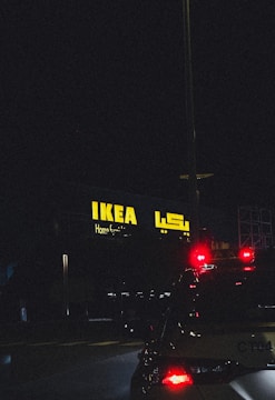 A night scene featuring an illuminated IKEA store sign in yellow with Arabic script next to it. The foreground features the rear of a vehicle with its brake lights glowing red, adding contrast against the dark surroundings.