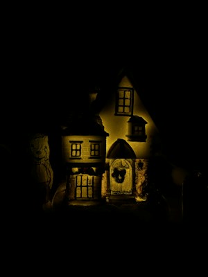 A dimly lit miniature model of a house with detailed features like windows and a door. The model is illuminated by a soft yellow light, which contrasts with the dark background, creating a mysterious atmosphere.